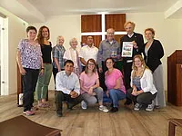 Gruppe von elf Personen posiert drinnen, einige halten einen gerahmten Gegenstand.