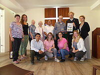 Gruppe von elf Personen posiert drinnen, einige halten einen gerahmten Gegenstand.