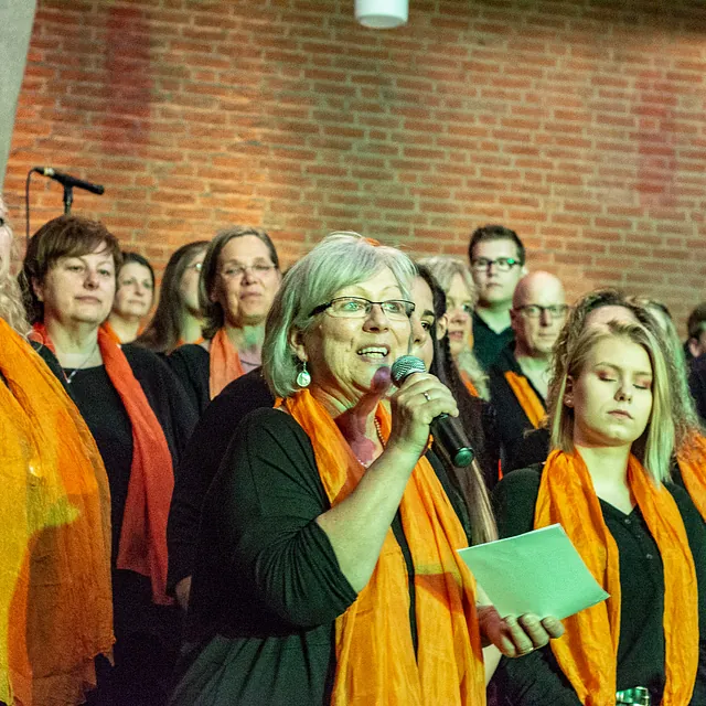 Chorauftritt mit Leadsängerin und orangen Schals, vielfältige Gruppe.
