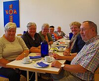 Senioren treffen sich an Tischen, genießen Getränke, Kreuz an der Wand.