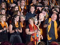 Großer Chor, verschiedene Altersgruppen, in Schwarz und Orange auftretend.