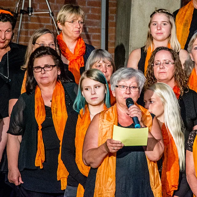 Frau mit orangem Schal singt ins Mikrofon, umgeben von Menschen.