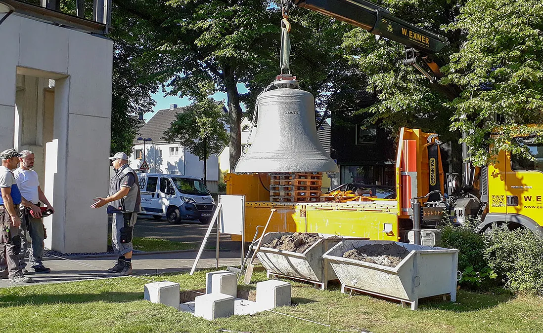 Große Glocke wird von Kran installiert, Arbeiter schauen zu.