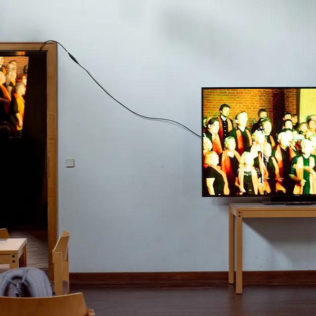 Fernseher zeigt Choraufführung, Tür zu anderem Raum mit Personen.