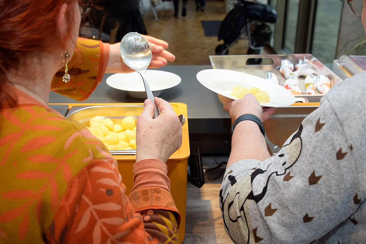 Zwei Personen bedienen sich an einem Buffet, Kartoffeln sichtbar.