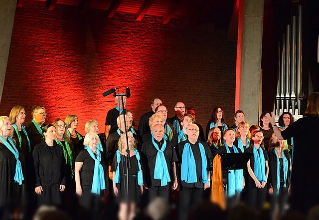 Chor in blauen und grünen Schals tritt auf, Dirigent im Vordergrund, rote Beleuchtung.