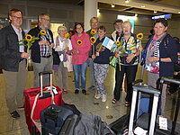 Gruppe Menschen mit Sonnenblumen und Gepäck am Flughafen, möglicherweise zur Begrüßung.