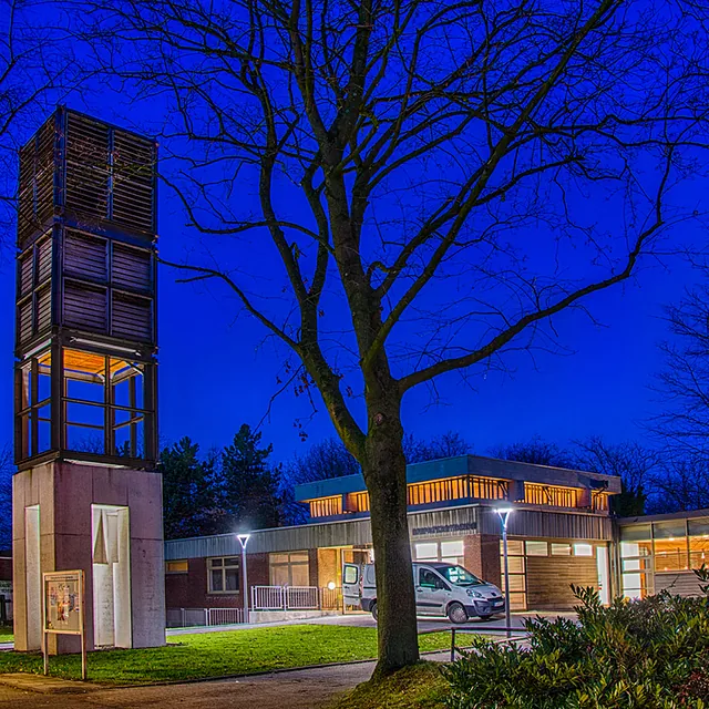 Moderner Kirchenkomplex in der Dämmerung, dunkelblauer Himmel, kahle Bäume.