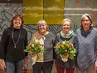 Vier lächelnde Frauen, zwei mit Sträußen, stehen drinnen vor Kunstwerken.