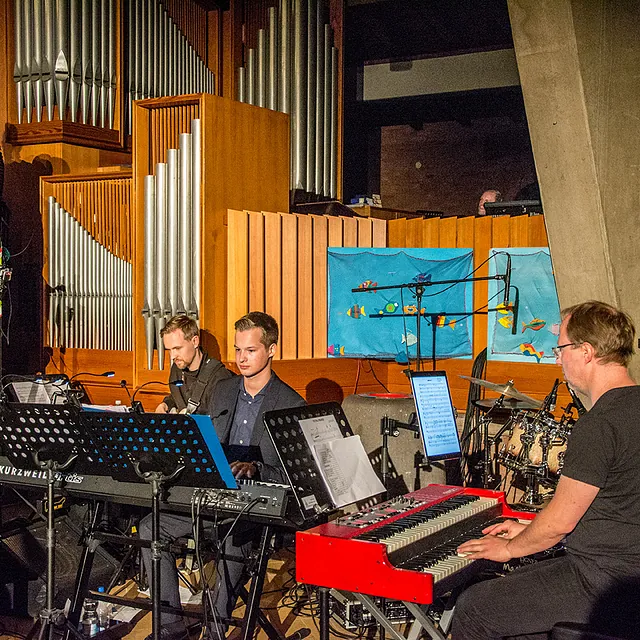 Musiker spielen Keyboards und Schlagzeug mit Chor daneben.
