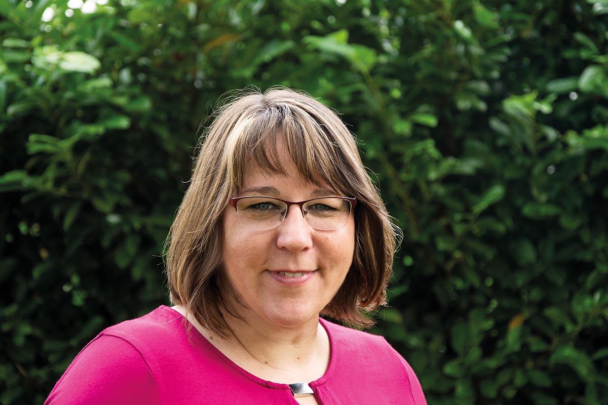 Eine lächelnde Frau mit Brille und pinkem Hemd vor grünem Laub.