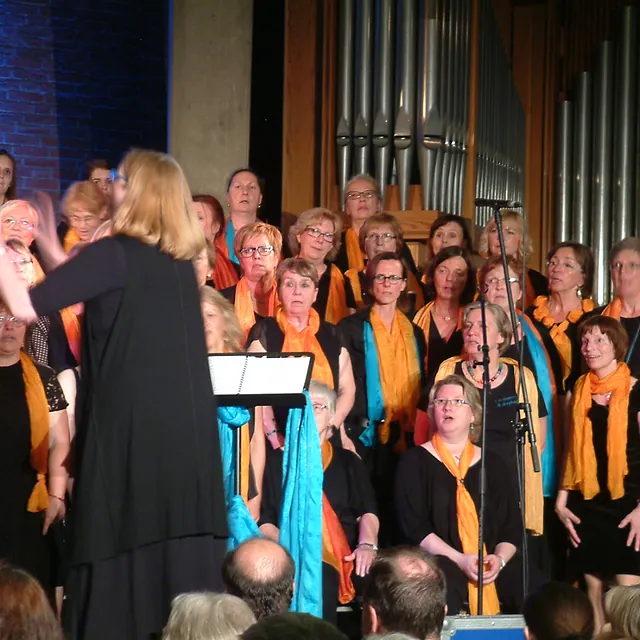 Chorleiterin leitet Chor, viele Mitglieder tragen Orange.