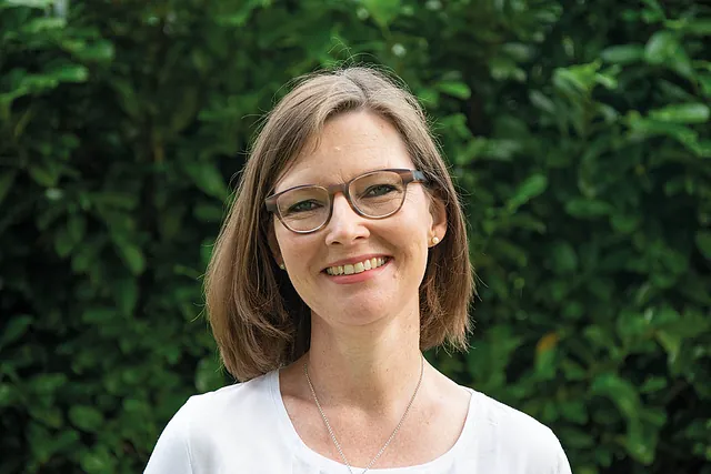 Eine lächelnde Frau mit Brille und weißem Hemd vor grünem Laub.