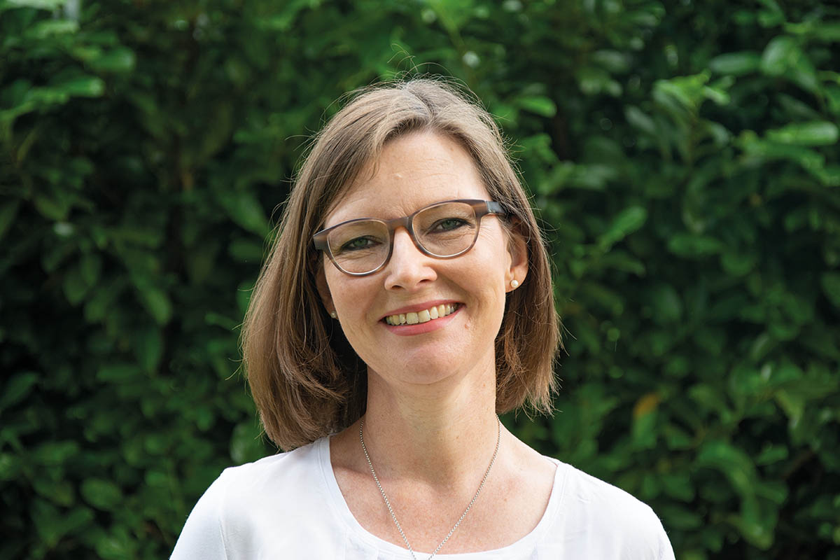 Eine lächelnde Frau mit Brille und weißem Hemd vor grünem Laub.