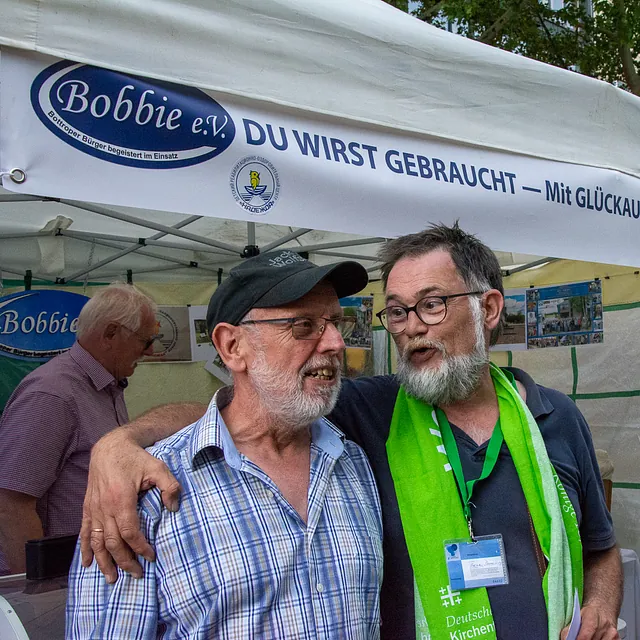 Zwei bärtige Männer umarmen sich lächelnd unter einem Zelt.