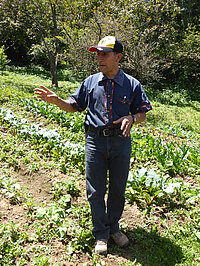 Ein Mann im Feld gestikuliert, über Pflanzen oder Landwirtschaft sprechend.