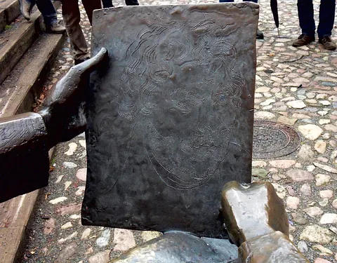 Bronzeskulptur: Hände halten Tafel mit geätztem Männergesicht.