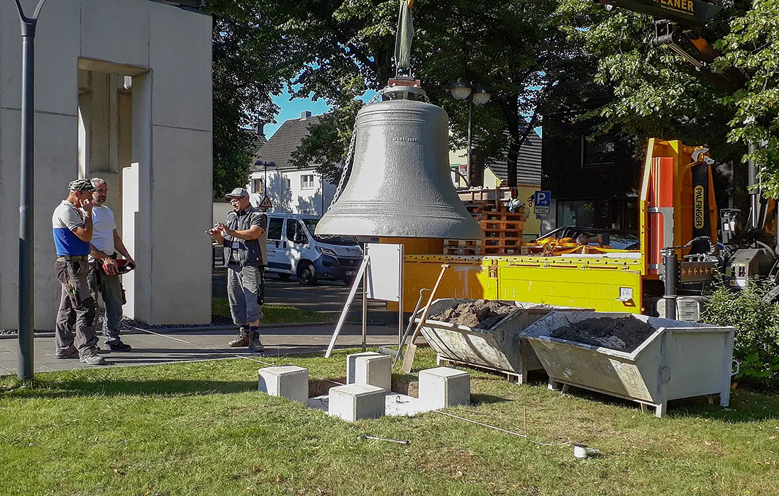 Große Glocke hängt am Kran während der Installation; Arbeiter beobachten.