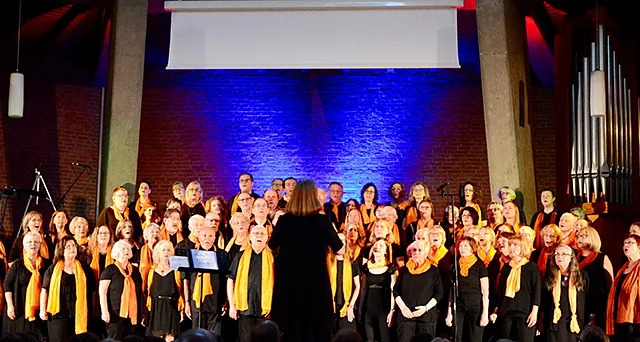 Großer Gospelchor auf der Bühne, Dirigent vor vielfältigen Sängern, blaue Beleuchtung.