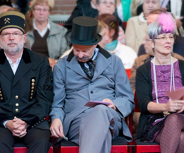 Drei Personen in historischen Kostümen sitzend, zwei schauen nach vorne.