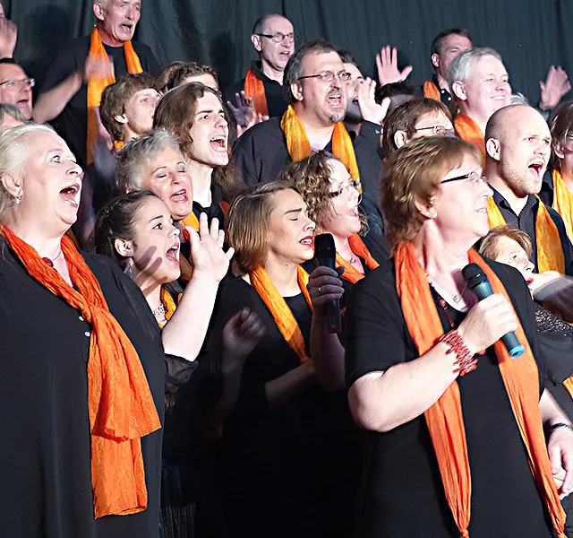 Begeisterter Chor singt mit offenem Mund, trägt orangefarbene Schals.