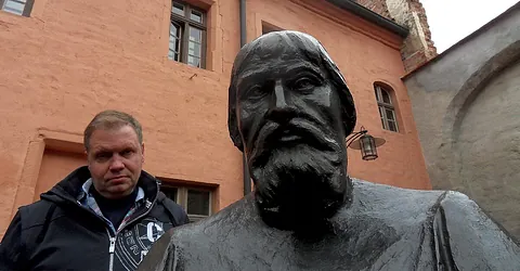 Mann steht neben dunkler, verwitterter Büste im Freien.