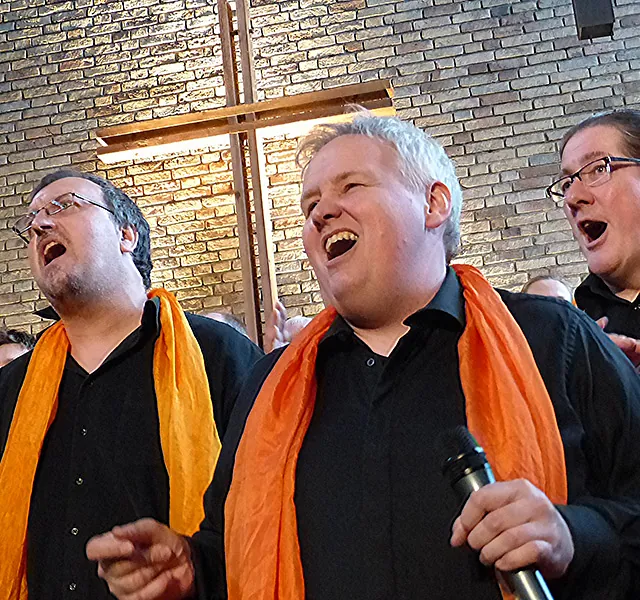 Drei Männer in schwarzen Hemden und orangefarbenen Schals singen.