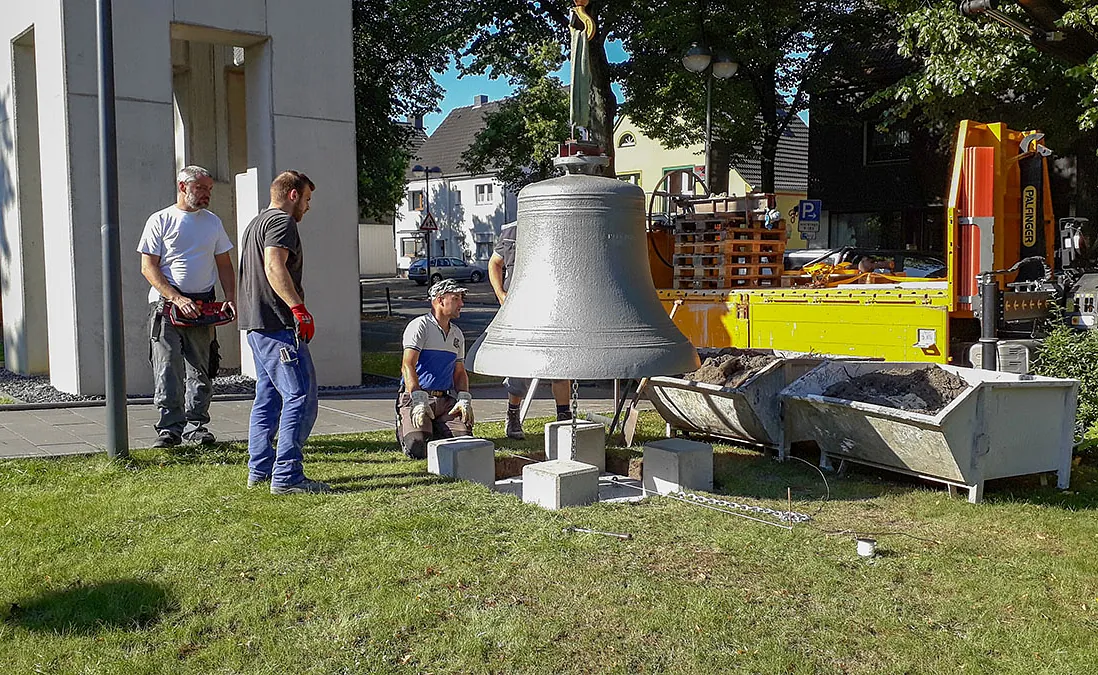 Arbeiter knien um eine große Glocke, die am Kran hängt.