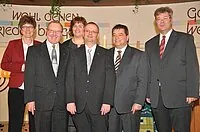 Gruppe von sechs Erwachsenen in Anzügen, teils lächelnd, drinnen posierend.