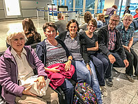 Sechs Erwachsene sitzen auf Bänken in einem Flughafen-Wartebereich.