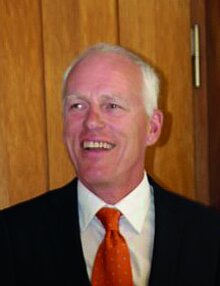 Lächelnder Mann mit kurzen weißen Haaren, dunklem Anzug, orangefarbener Krawatte.