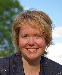 Lächelnde Frau mit kurzen blonden Haaren, blauen Augen und Sommersprossen.